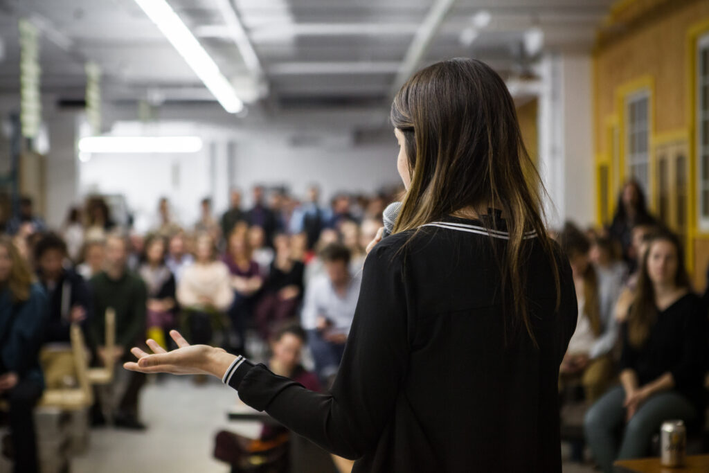A woman speaking into a microphone to a large, attentive audience in an open workspace.