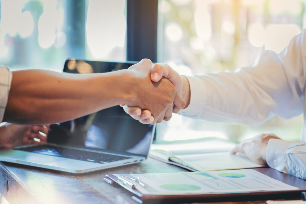 Two professionals shaking hands over a desk with a laptop and financial charts.