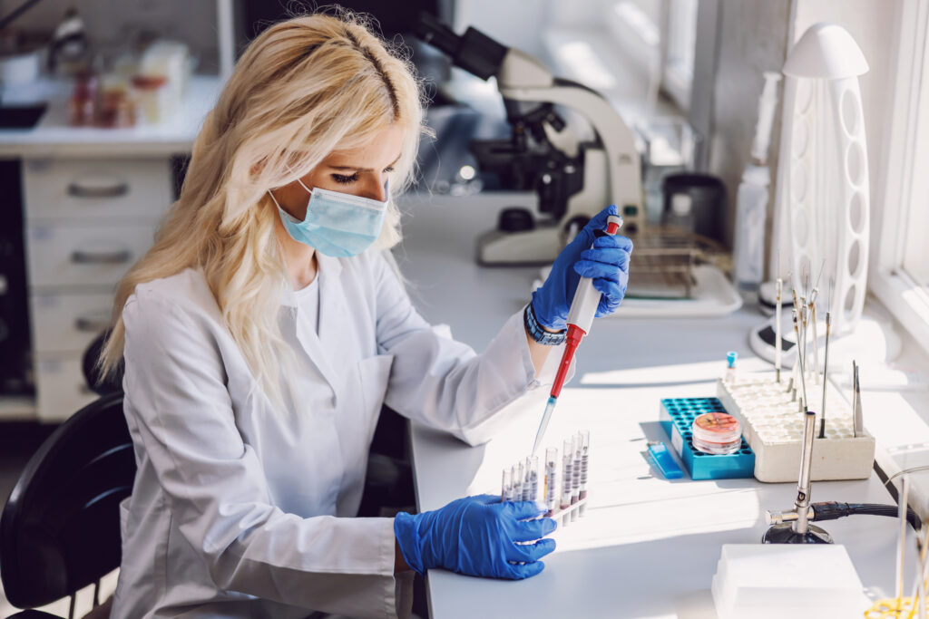 A female scientist in a lab coat and mask using a pipette in a laboratory.