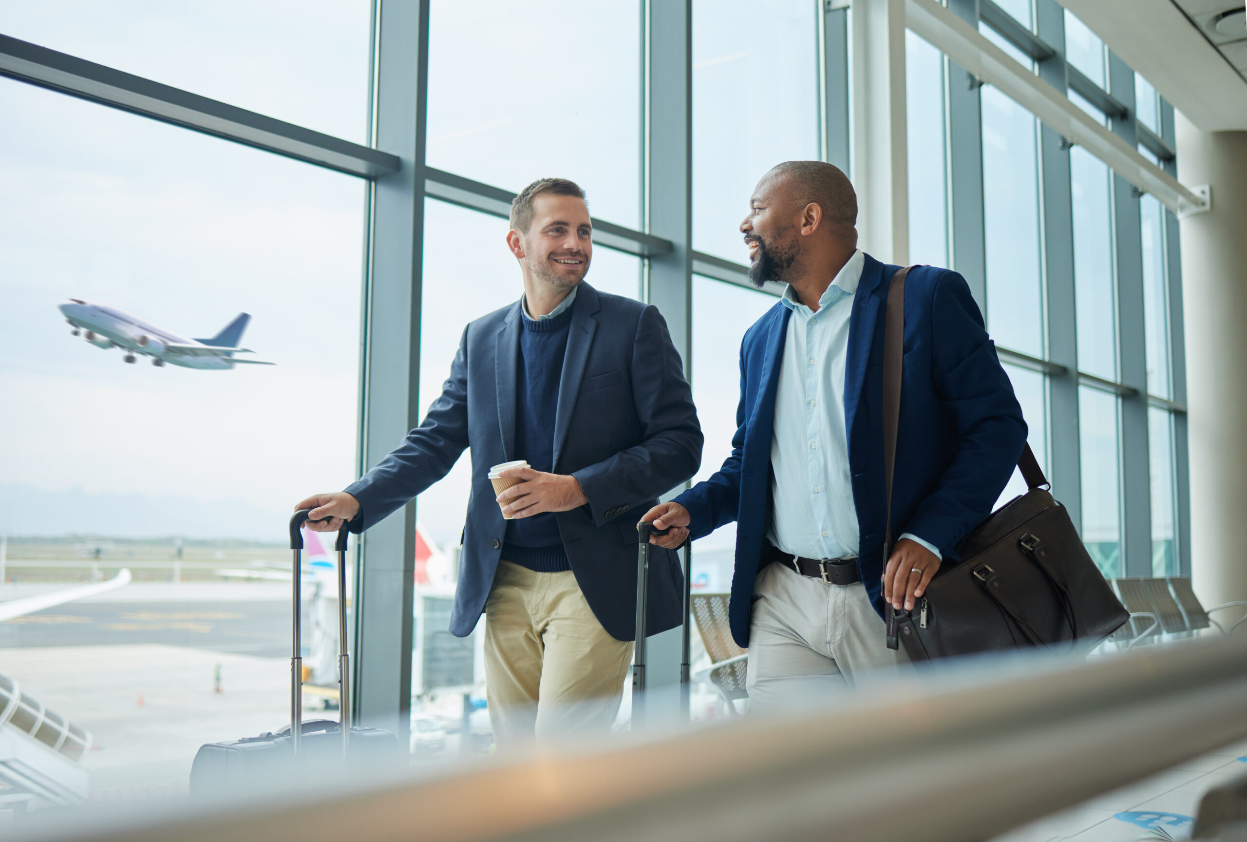 Two men in professional attire walking through an airport terminal with rolling luggage.
